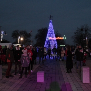 grupa ludzi wieczorem na zewnątrz a za nimi wielka choinka ze światełek i napis „Wesołych Świąt".