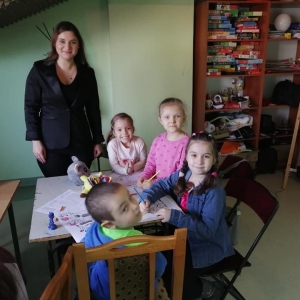 Pani Dyrektor Paulina Frontczak-Pawłowska stoi uśmiechnięta obok dzieci kolorujących przy stole obrazki.