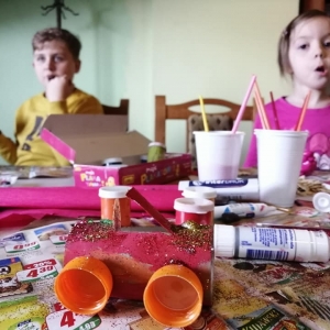 Dzieci siedzą przy stole pełnym przyborów plastycznych.