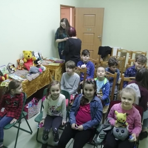 Dzieci siedzą na ustawionych rzędami krzesłach. 